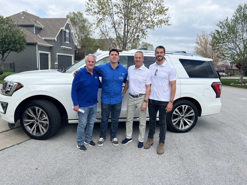 A family run business, four men all related, standing in front of the car in a neighborhood.